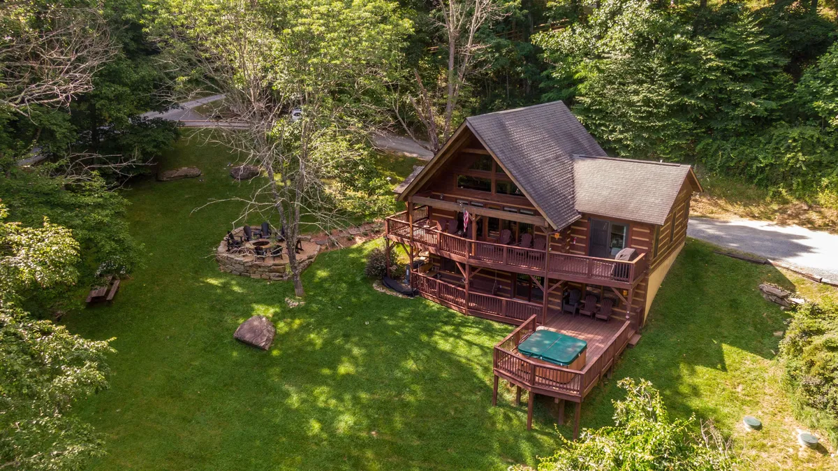 A welcoming view of the cabin’s exterior shows the spacious hot tub and a stone fire pit nestled among the surrounding trees. The log cabin and outdoor features are integrated into the natural landscape, creating a serene atmosphere ideal for relaxation and gatherings.