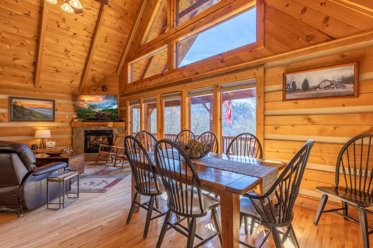 A spacious dining and living area with floor-to-ceiling windows revealing views of the natural surroundings. The area includes a large dining table, comfortable seating, a fireplace, and decorative touches, all bathed in natural light from the expansive windows.