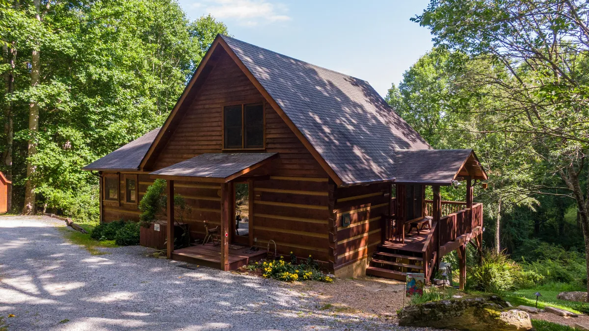 A cozy log cabin with an inviting wooden deck and vibrant flower beds, nestled in a serene natural setting. The deck and porch areas provide ample outdoor space for pets and owners alike to enjoy the mountain environment.