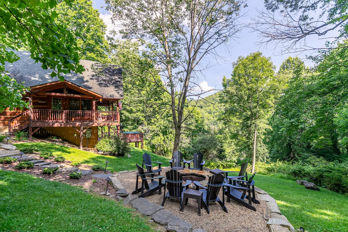The focal point is a cozy fire pit encircled by Adirondack chairs, backed by wooded surroundings and the rustic log cabin. The setup invites guests to enjoy social evenings, s’mores, and stories in a natural setting.