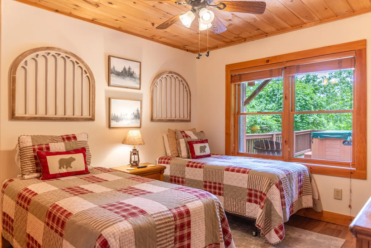 A bedroom with two twin beds dressed in plaid bedding, a wooden ceiling, a large window offering outdoor views, and decorative wall art. The space feels bright and comfortable, ideal for relaxing or sharing with friends or family.