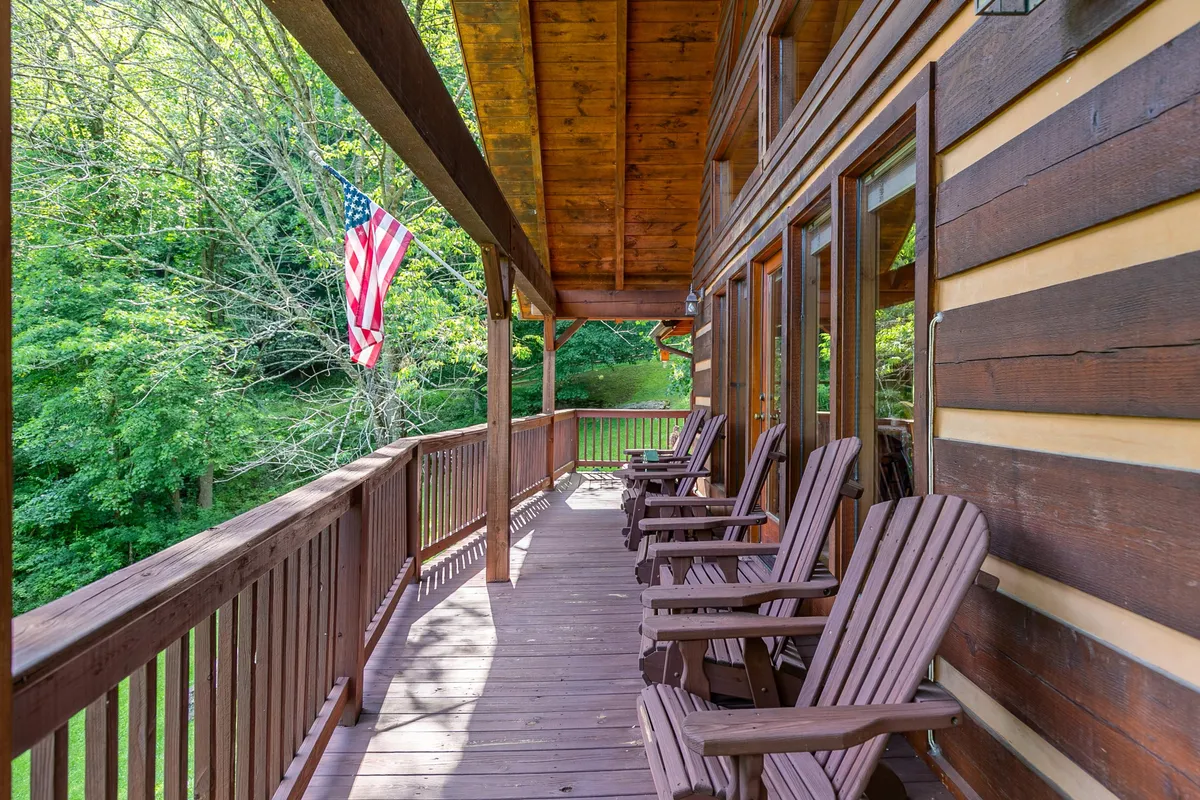 This exterior image features the cabin’s inviting front porch with classic wooden rocking chairs and an American flag gently waving in the mountain air. The wooden railing frames a view of the surrounding natural landscape, suggesting a peaceful retreat that invites guests to socialize and enjoy the outdoors together.