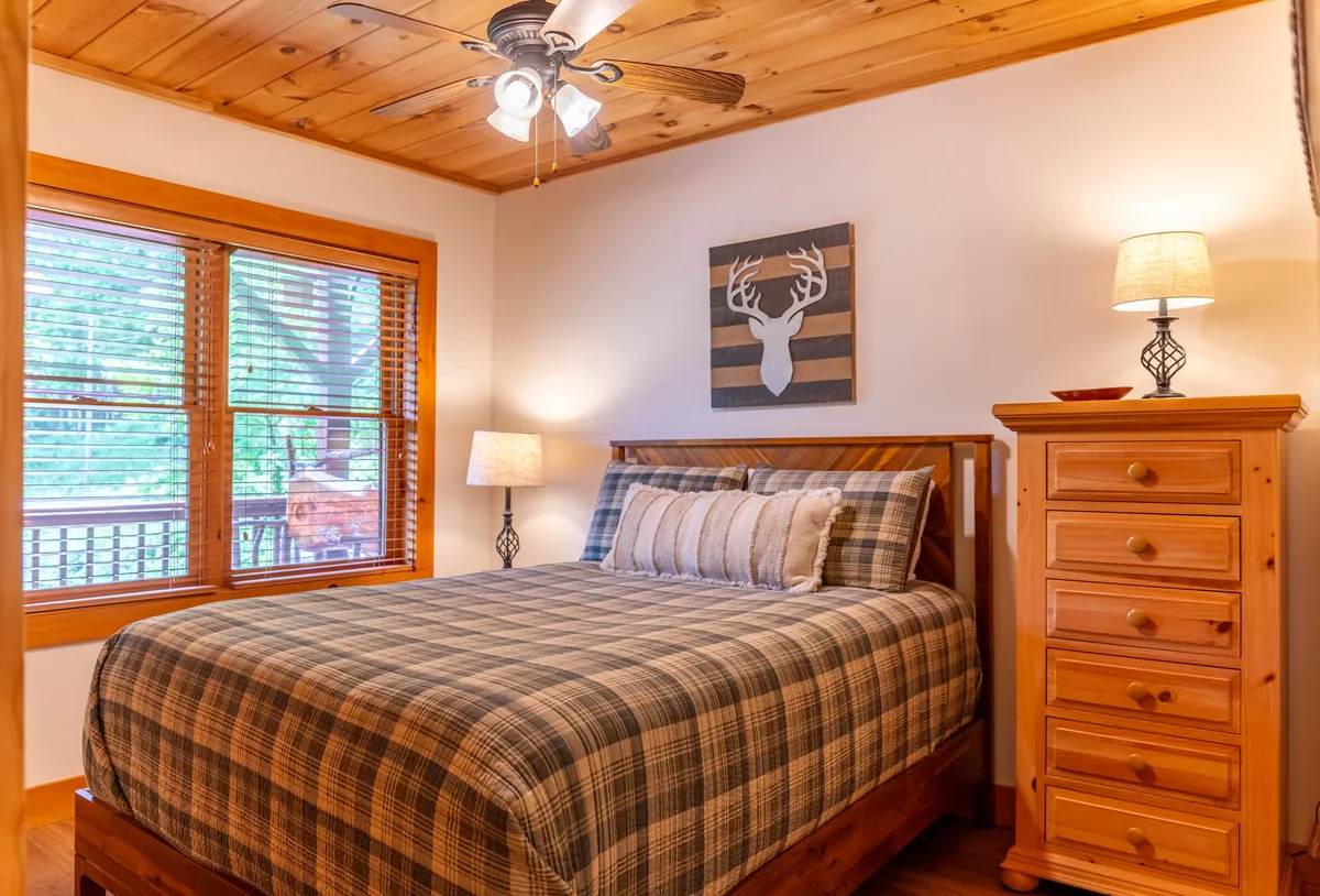 A cozy bedroom featuring a bed with plaid bedding, a wooden chest of drawers, a bedside table with lamp, and framed deer artwork above the bed. Soft natural light brightens the space, emphasizing warmth and tranquility.
