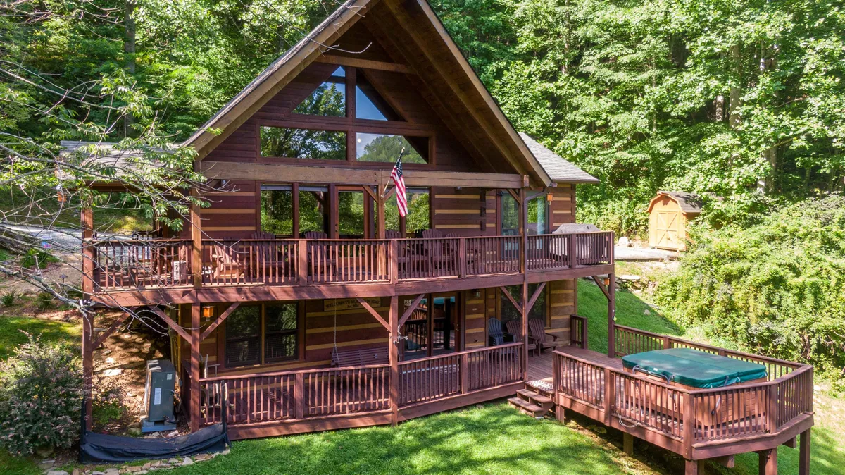 A broad wooden deck featuring a hot tub, large windows looking into the cabin, and a visible flag. The deck offers open space for relaxation, barbecues, and is an easy-access area for guests and their pets.