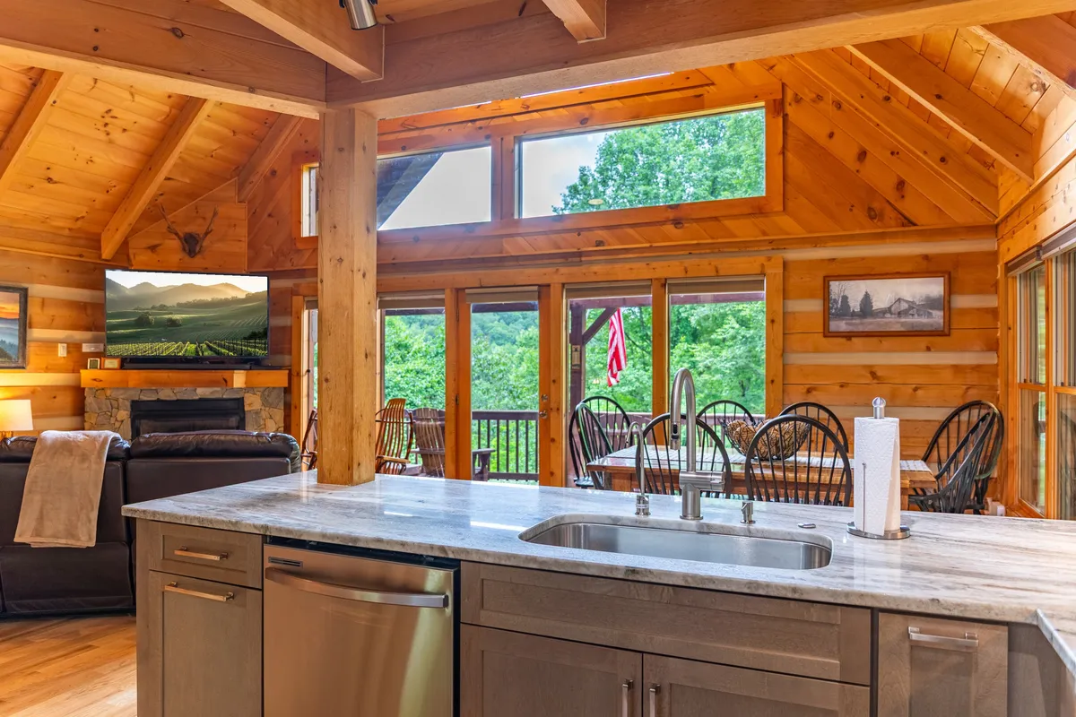 A spacious open-concept living space that seamlessly connects the kitchen, living area with a TV, dining area, and large windows framing beautiful outdoor views. The kitchen island and adjacent living room are perfect for entertaining and gathering.