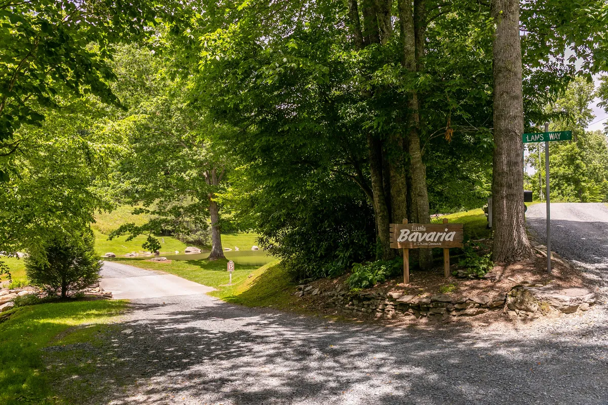 A gravel road bordered by green foliage leads up to a sign marked 'Bavaria,' signaling entry into the private community. Stone edging and idyllic greenery set a calming, welcoming tone, showcasing the exclusivity and natural beauty of the area.