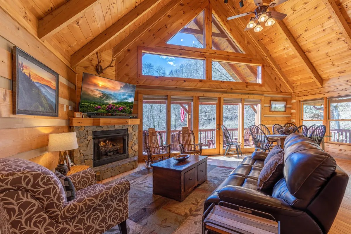 A warm and inviting family room featuring a comfortable seating area oriented around a stone fireplace and a large TV, with expansive windows that offer views of the surrounding nature. The dining table is positioned to one side, inviting family meals and gatherings.
