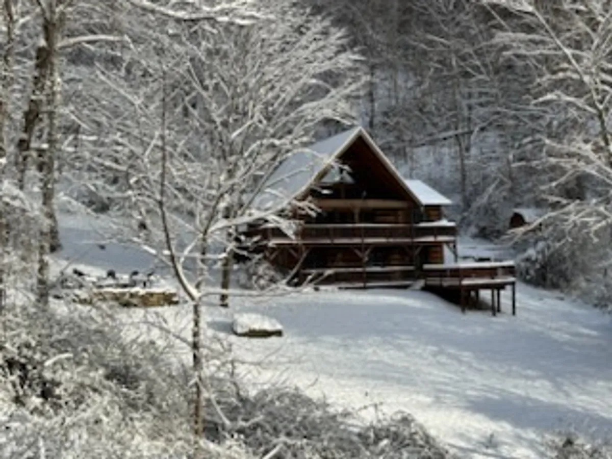 A log cabin set against a snow-covered landscape with bare winter trees. The cabin features a wrap-around porch, and the surrounding scenery emphasizes the property’s mountain setting and serene atmosphere.
