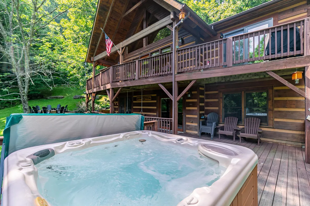 A steaming hot tub sits on a spacious wooden deck, surrounded by lush greenery and attached to a classic log cabin structure. The deck offers plenty of room for guests to lounge, socialize, or enjoy a barbecue while immersed in the sights and sounds of nature.