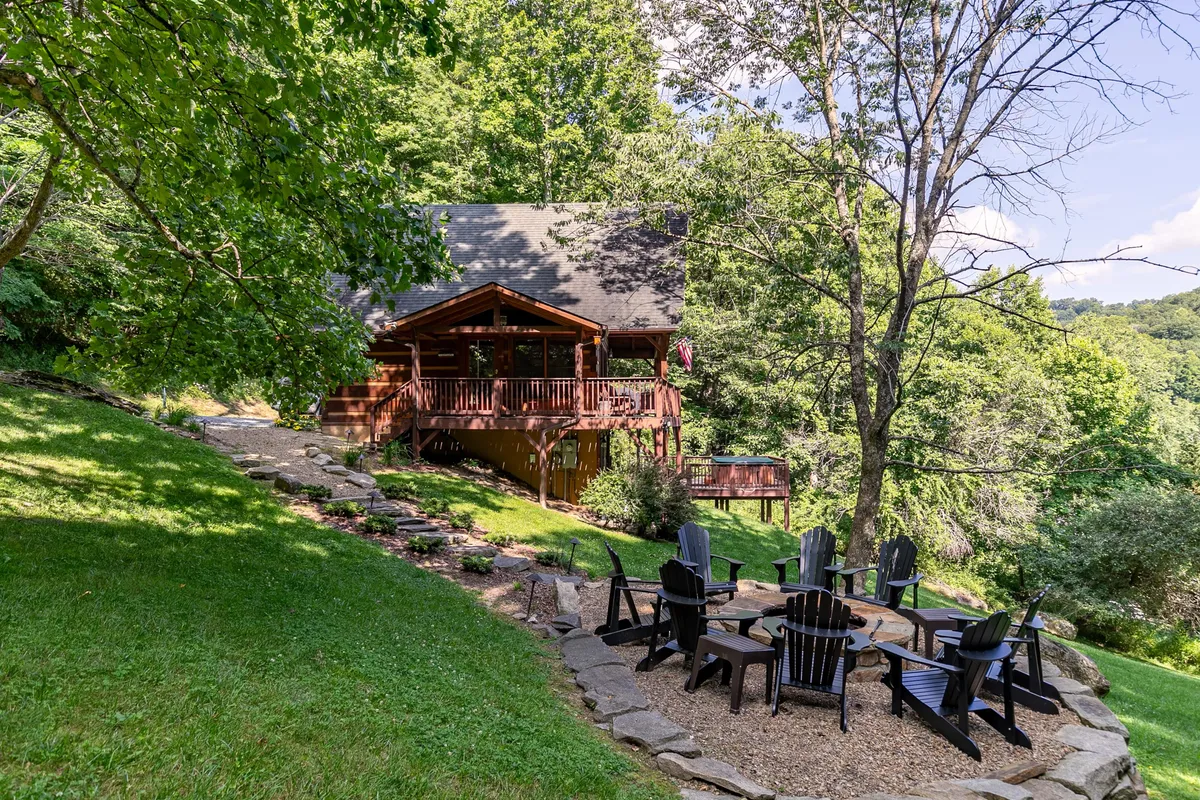 A log cabin set among towering trees with an inviting outdoor seating area and a stone pathway leading to a fire pit. The deck offers views of the sunset, and the setting is ideal for family gatherings and outdoor activities like roasting s'mores or enjoying the mountain air.