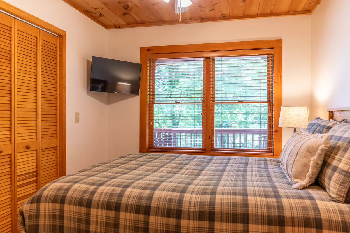 A bedroom with a plaid bedspread, large windows allowing in natural light, wooden furniture, a mounted TV, and a lamp. The cozy setting invites relaxation, TV watching, and enjoying the view outside.