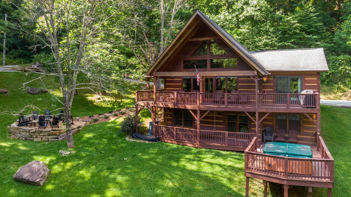 The image showcases a charming log cabin nestled in the North Carolina mountains, featuring expansive decks, a bubbling hot tub, and a cozy fire pit area. The outdoor space is set up for relaxation and gatherings, with views of rolling hills and lush forests in the background, creating a tranquil and inviting atmosphere.