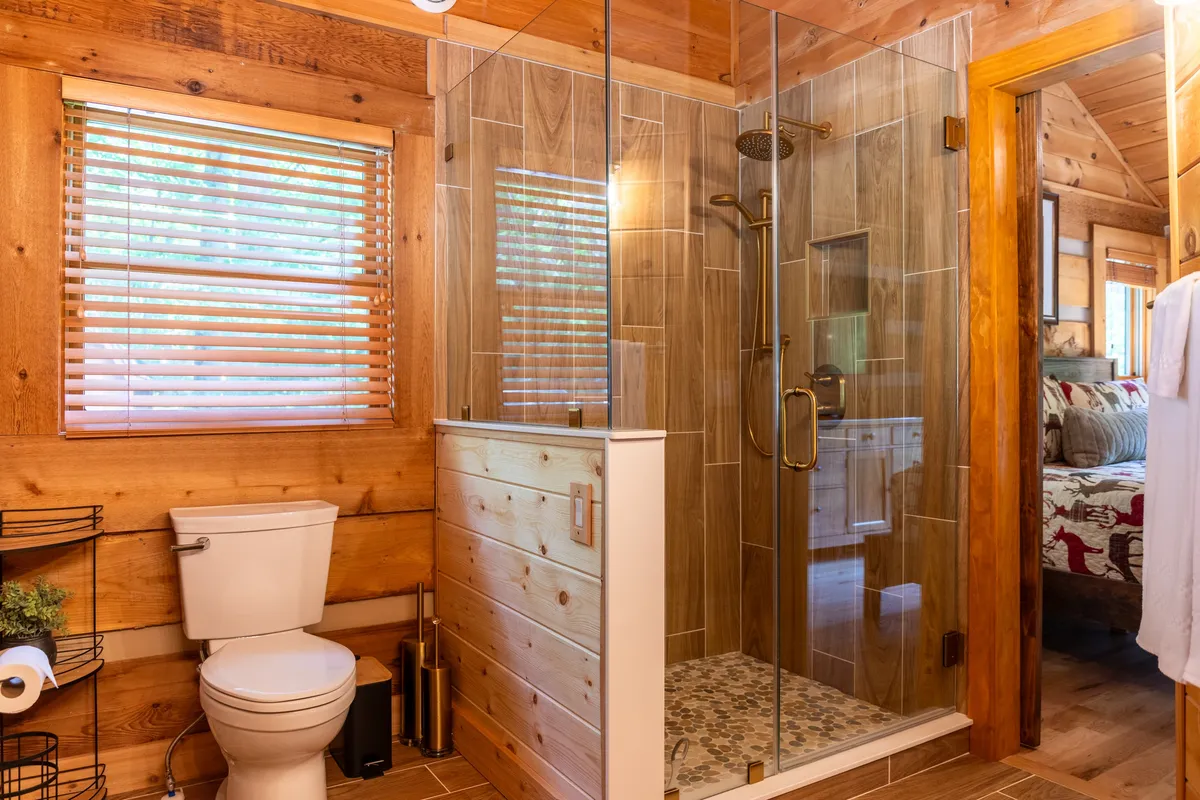 A modern bathroom with a glass shower enclosure, wooden accents, decorative tile flooring, and a window with blinds. The scene is serene and comfortable, suggesting a spa-like atmosphere ideal for relaxation.