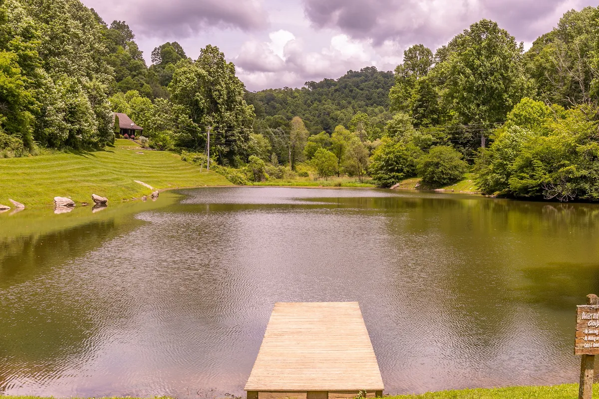 A tranquil private lake surrounded by lush trees, with a wooden dock extending over calm water. The distant cabin and early morning mist evoke a serene, secluded atmosphere ideal for fishing, boating, or peaceful reflection. Wildlife can sometimes be spotted along the banks, and the sunset reflections create a magical effect.